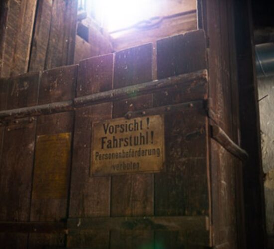 Das Foto zeigt einen alten Fahrstuhl aus Holz. Man kann das Schild erkennen "Vorsicht! Fahrstuhl! Personenbeförderung verboten."